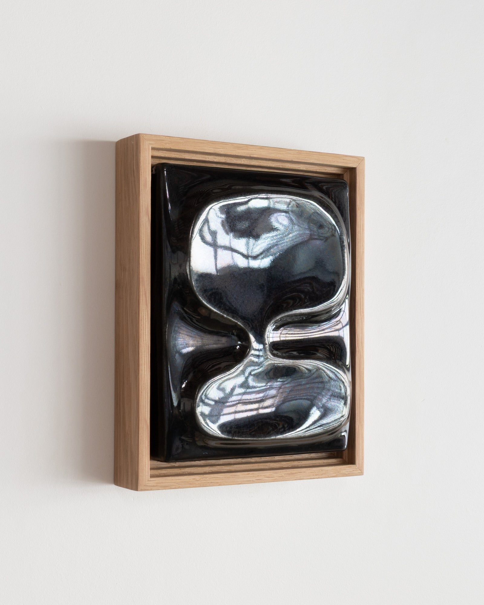Angled view of a small black reflective ceramic wall relief in a wooden frame